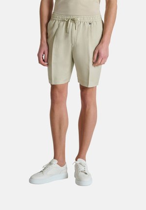 Muškarac u bež šortsevima s elastičnim pojasom i vezicama te bijelim tenisicama, stoji ispred jednobojne pozadine.