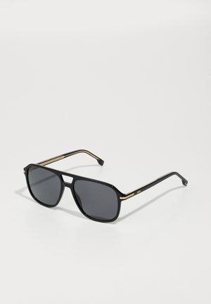 Black sunglasses with a square frame, grey lenses, gold accents on the temple, and a glossy finish. Distinctive cut at the bridge.