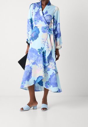 Kvinde iført en blå blomsterprintet omslagskjole med lange ærmer og flæset asymmetrisk kant, holder en sort clutch og har på lyseblå sandaler med hæl.