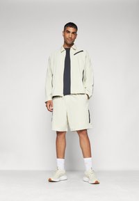 Light beige athletic outfit featuring a zip-up jacket with black accents, a black shirt, and matching shorts. Sneakers with textured soles.