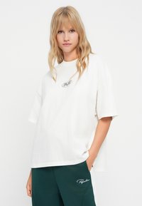 Tricou alb din bumbac cu mâneci scurte, având un logo verde circular în centru. Asortat cu pantaloni de jogging verde închis cu accente albe.