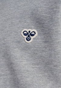 Marineblåt og hvidt broderet bi-logo på lys grå strikket stofbaggrund.