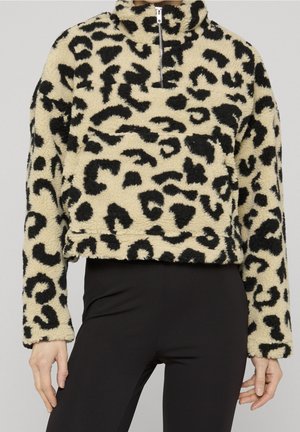 Pull en polaire beige et noir à imprimé léopard, avec demi-fermeture éclair et manches longues, porté avec un pantalon noir, vue de face de la taille vers le haut.