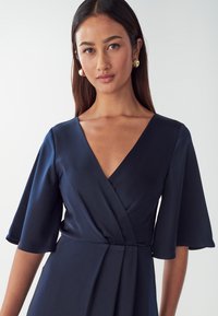 Navy blue wrap dress with a deep V-neck, short flared sleeves, and a smooth satin texture. Features a fitted waist and pleated skirt.