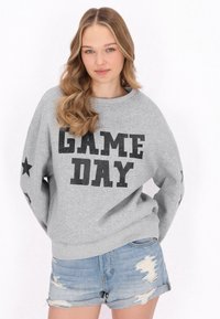 Felpa grigia con la scritta "GAME DAY" in caratteri neri e in grassetto, con stelle nere sui maniche e una vestibilità rilassata. Indossata con pantaloni shorts in denim.