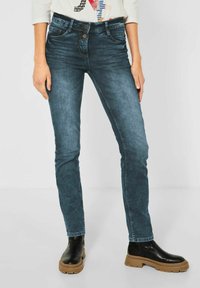 Jeans de mezclilla azul oscuro con un ajuste slim, que cuentan con dos bolsillos frontales, cierre de botón y una textura ligeramente descolorida. Combinados con botas negras.