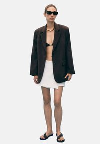 Blazer noir oversize, haut de bikini noir, mini-jupe blanche, lunettes de soleil et tongs. Présente un design structuré et des textures variées.