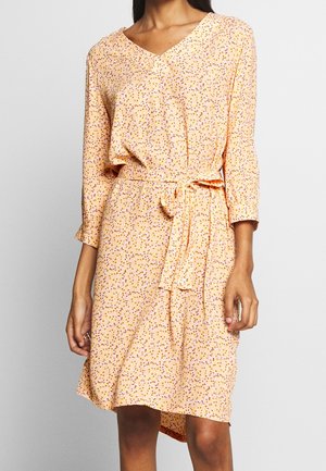 Woman wearing a light peach midi dress with 3/4 sleeves, small multicolor floral print, V-neckline, and a tied waist belt.