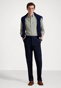 Granatowa dzianinowa kamizelka na koszuli w kratę z długim rękawem, zestawiona z ciemnymi spodniami i brązowymi butami. Okulary i zegarek dopełniają strój.