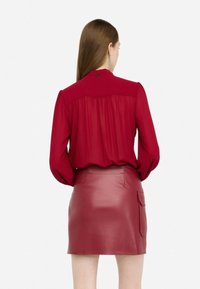 Blusa rossa con schiena arricciata e maniche lunghe, abbinata a una mini gonna in pelle cremisi attillata con tasca laterale.