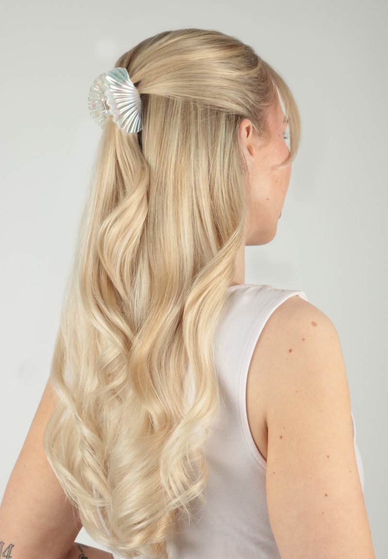 Femme blonde aux cheveux longs et ondulés, mi-attachés à l'arrière avec une barrette brillante en forme de coquillage, portant un haut blanc sans manches.