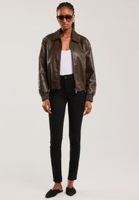 Naine, kes kannab tumepruuni nahktagi, valget pluusi, musti pükse, musti libisevaid kingi ja päikeseprille, seisab ühevärvilise heleda tausta ees.