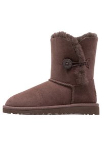 Brązowy zamszowy botek za kostkę z okrągłym noskiem, ozdobnym guzikiem, sztucznym futrem w środku i teksturowaną gumową podeszwą. Płaski krój, luźny fason.