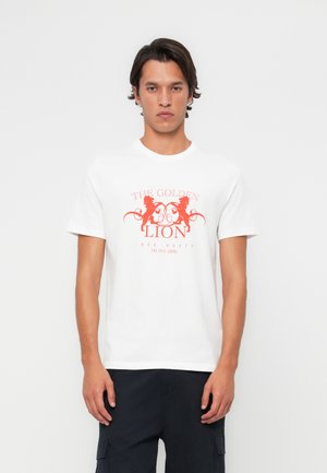 T-shirt blanc à manches courtes avec une impression graphique rouge représentant "LE LION D'OR" et des motifs ornés. Fabriqué en matériau en coton doux.