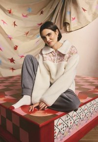 Hanorac alb din fleece cu panou și guler cu model floral, însoțit de pantaloni de trening gri. Fundal în carouri roșii și roz cu elemente decorative.