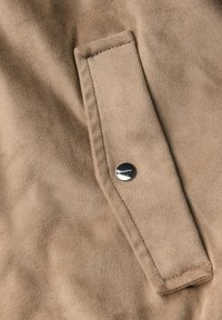 Tessuto scamosciato marrone chiaro con tasca a patta dotata di chiusura a scatto metallico, che mostra una texture liscia e una leggera lucentezza.