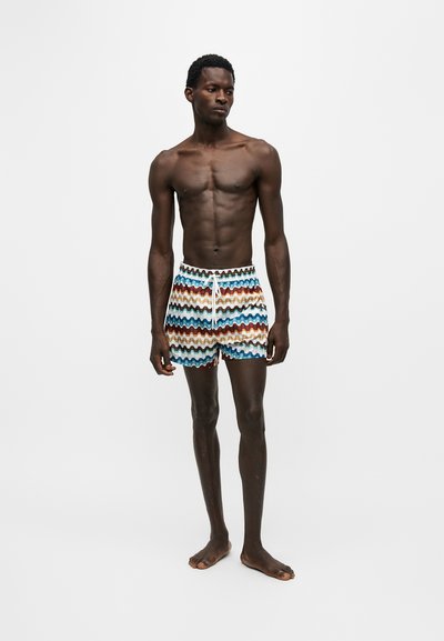 Homme athlétique à la peau foncée, debout pieds nus, portant un short de bain rayé ondulé multicolore avec un cordon blanc, sur un fond clair uni.