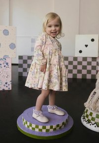 Lyserød kjole med blomsterprint, lange ærmer og lagdelt nederdel. Hvide sko med ankelsnørebånd, stående på en farverig piedestal.