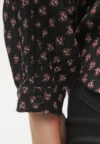 Blouse noire avec de petits motifs floraux en rose et blanc. Dispose de manches froncées et d'une fermeture à bouton au niveau des poignets. Tissu léger.