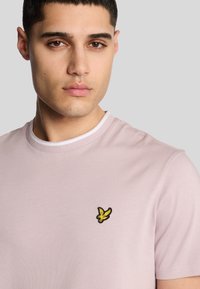 Lys pink bomulds-t-shirt med rund hals og korte ærmer, prydet med et lille broderet sort og gult fuglelogo på brystet.