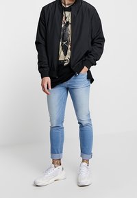 Czarna kurtka bomberka, pod nią koszulka z nadrukiem, jasnoniebieskie wąskie dżinsy, białe trampki z akcentami oraz widoczne tatuaże na kostkach.