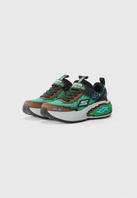 Een paar sneakers met een pixelthema in groen, bruin en zwart, voorzien van klittenband, veters en een dikke, gemusterde zool op een witte achtergrond.