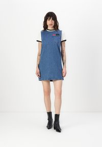 Vestido de ganga azul claro com design sem mangas, bolso frontal e detalhe na barra. Combinado com uma camisa de manga curta preta e branca e botins pretos. 