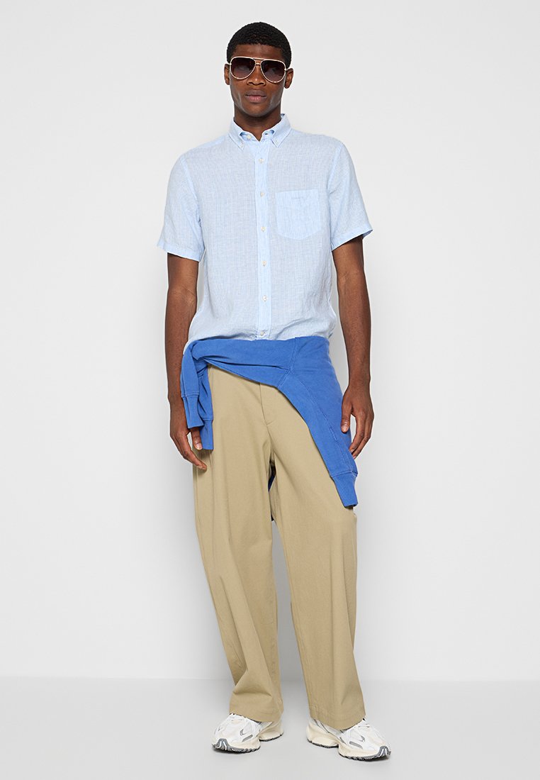 Kurzarm, hellblaues Shirt, khakifarbene Hose, blauer Pullover um die Taille gebunden und Sonnenbrille. Glattes Material, lockere Passform, sportlicher Stil.