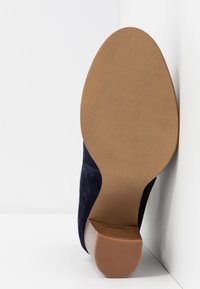 Semelle d'une botte cheville en daim bleu marine avec un dessous en caoutchouc lisse couleur tan et un talon bloc en bois empilé sur fond blanc.
