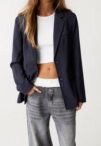 Veste bleu marine à deux boutons, coupe ajustée, avec un revers cranté, associée à un top blanc court et à un jean gris taille haute.