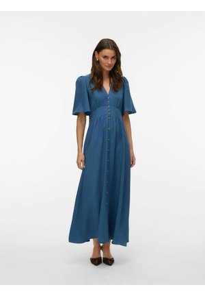 Kvinne står iført en blå midi-kjole med knapper og korte, puffete ermer, og svarte spisse flate sko, mot en hvit bakgrunn.