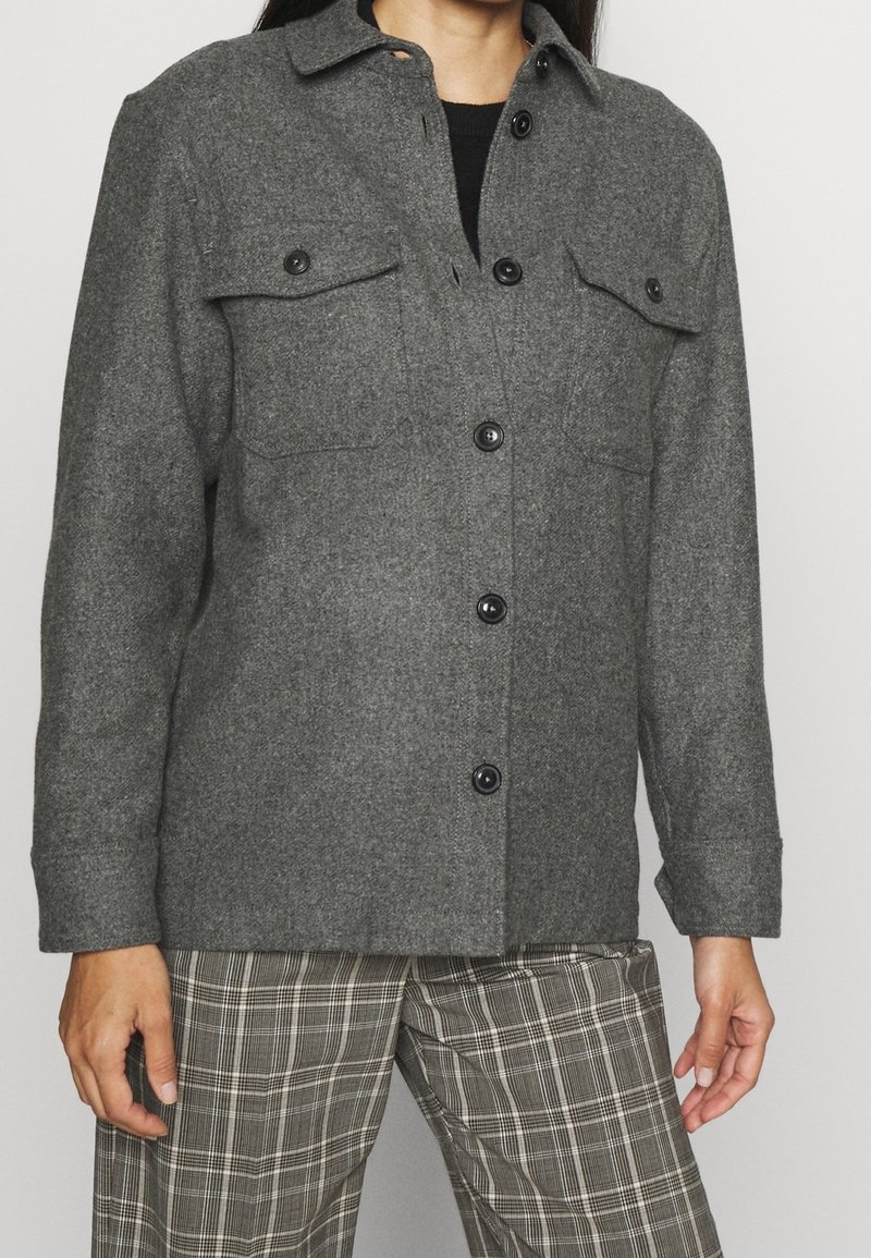 Giacca camicia in lana grigia con bottoni neri, due tasche sul petto e colletto, abbinata a pantaloni a quadri fantasia.