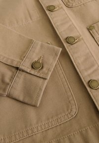 Veste en toile beige avec une surface texturée, des boutons en métal et des coutures apparentes. Comprend des poches à rabat et un col structuré.