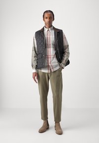 Colete acolchoado preto sobre uma camisa xadrez com padrões em cinza, branco e vermelho. Calças verde-oliva e botins bege completam o visual.