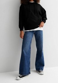 Sudadera con capucha negra y bolsillo canguro, combinada con vaqueros de denim azul de pierna ancha y zapatillas negras. La textura es suave y casual.