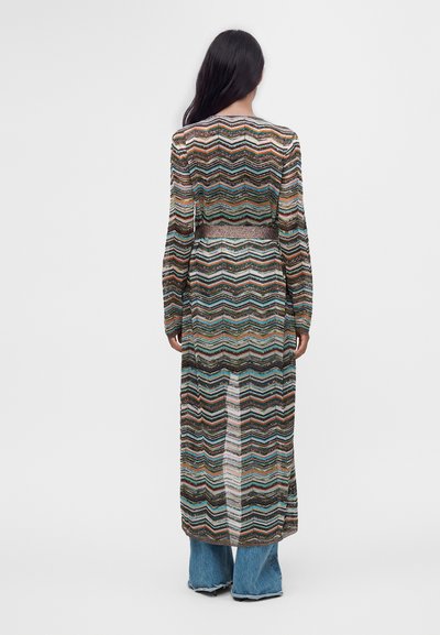 Femme aux longs cheveux foncés portant un long cardigan à motif zigzag multicolore par-dessus un jean bleu, debout, de dos, sur un fond uni.