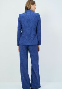 Costume bleu à motifs floraux en tissu texturé, comprenant une veste ajustée et un pantalon à jambes larges, présenté de dos.