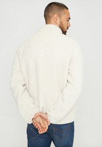 Giacca beige in pile con colletto alto e maniche lunghe. Il tessuto ha una consistenza morbida, con una vestibilità comoda e polsini leggermente elasticizzati.