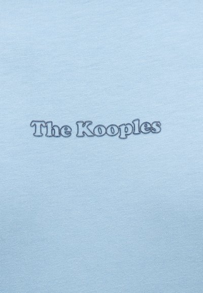 Ανοιχτό μπλε ύφασμα με κεντημένο το όνομα της μάρκας "The Kooples" σε σκούρο μπλε, τοποθετημένο στο κέντρο της επιφάνειας.