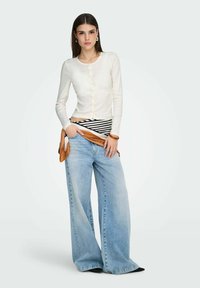 Cardigan beige clair à manches longues avec fermeture à boutons, associé à un jean large bleu clair. Les accessoires comprennent une écharpe marron nouée à la taille.