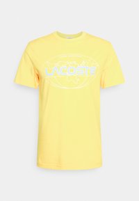 Žluté bavlněné tričko s krátkými rukávy, na němž je bílá grafika loga zahrnující světovou mapu a text "LACOSTE INTERNATIONAL."