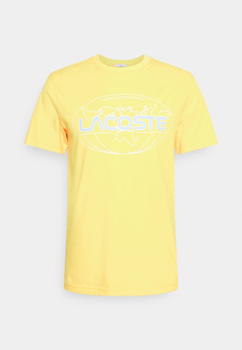 Žluté bavlněné tričko s krátkými rukávy, na němž je bílá grafika loga zahrnující světovou mapu a text "LACOSTE INTERNATIONAL."