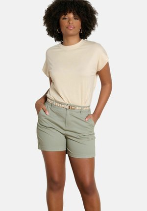 Vrouw met krullend haar, gekleed in een beige t-shirt, gevlochten riem en lichtgroene shorts, staand met handen in de zakken tegen een witte achtergrond.