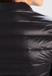 Personne portant une veste noire, brillante et matelassée, montrant le haut du dos et la zone du cou sur un fond neutre.