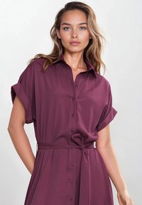 Bordowa sukienka koszulowa z lekkiego materiału, z przodem na guziki, krótkimi podwiniętymi rękawami i przewiązywanym w talii paskiem, co nadaje dopasowany krój.