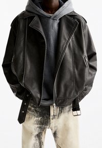 Personne portant une veste en cuir noir de motard sur un sweat à capuche gris et un jean clair éclaboussé de noir, les mains dans les poches de la veste.