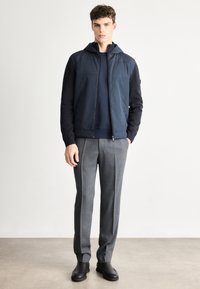 Jūras kara flotes tumši zils džemperis ar rāvējslēdzēju un kapuci, kontrastējošām piedurknēm, kopā ar pelēkiem piegrieztiem biksēm un melniem potīšu zābakiem. Gluda auduma tekstūra.