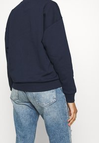 Mörk marinblå sweatshirt med ribbade muddar och nederkant, med en rundad halsdesign. Kombinerad med ljusblå slitna jeans.