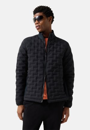 Young man wearing a black quilted jacket, orange shirt underneath, black pants, and dark sunglasses, standing against a white background.