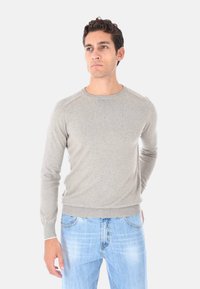 Uomo che indossa un maglione grigio chiaro a maniche lunghe e jeans azzurri chiari, in piedi contro uno sfondo bianco uniforme.